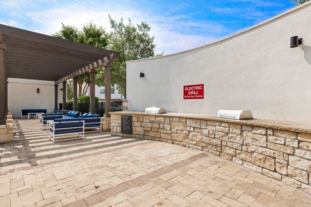 Outdoor patio area with blue lounge seating under a pergola, stone wall with two electric grills, and a red sign that says ELECTRIC GRILL. Trees and a blue sky are visible in the background.