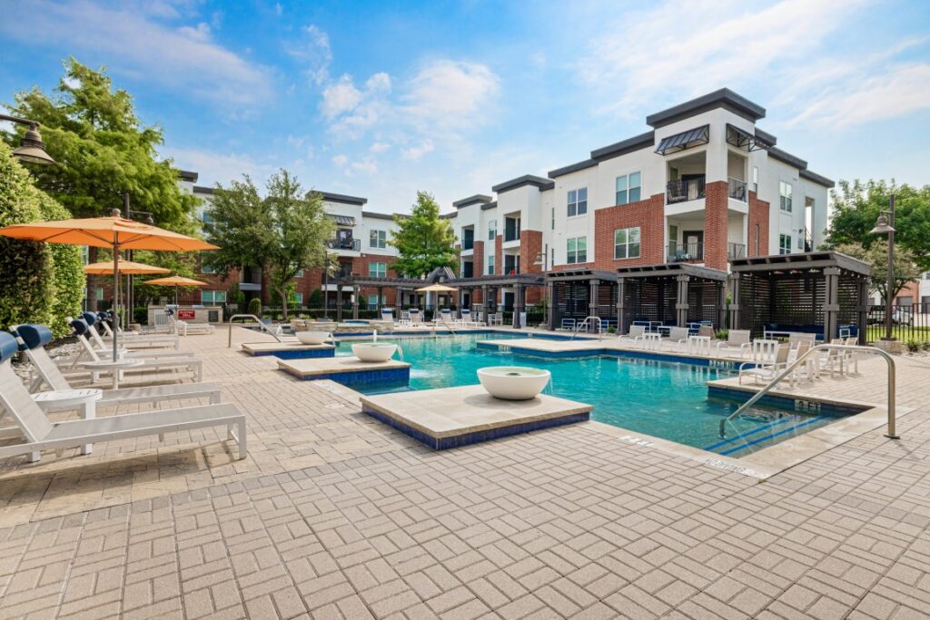Modern apartment complex with a large outdoor pool, lounge chairs, umbrellas, and landscaped trees under a blue sky with scattered clouds.