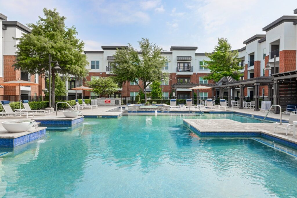 Modern apartment complex pool area with lounge chairs, umbrellas, and shaded seating. Trees and multi-story buildings with balconies surround the bright, inviting swimming pool under a blue sky.