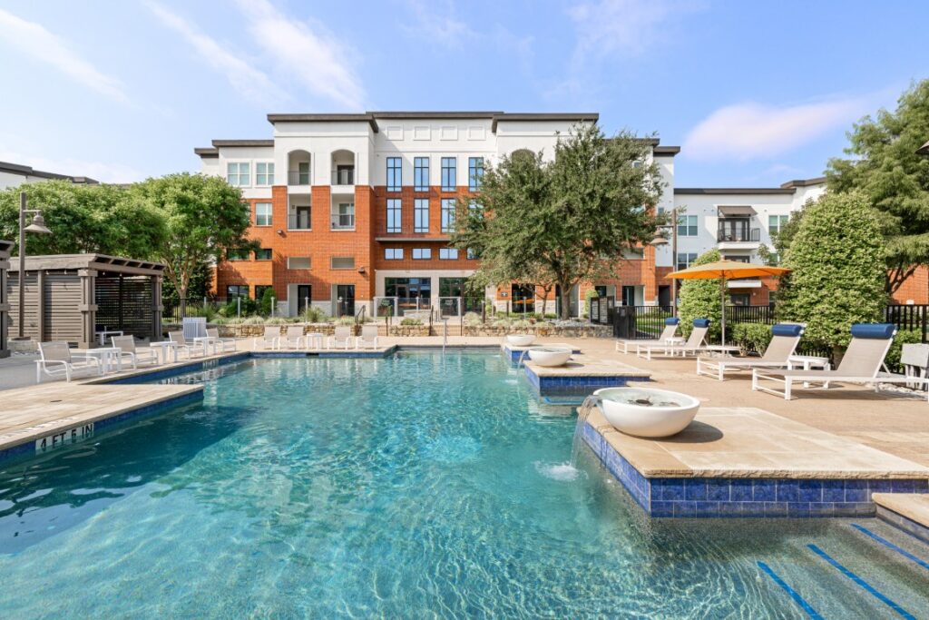 Modern apartment building with balconies behind a swimming pool, lounge chairs, umbrellas, and a shaded seating area, all surrounded by landscaping and clear blue sky.
