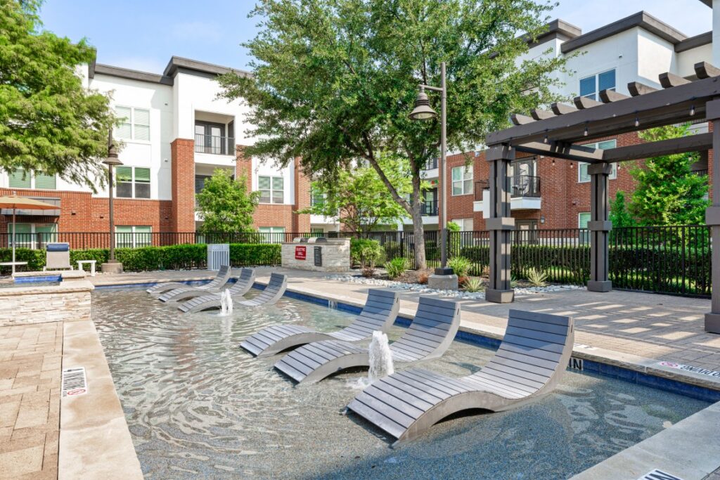 Shallow pool with built-in lounge chairs, small fountains, and a pergola. Modern apartment buildings and trees surround the pool area on a sunny day.