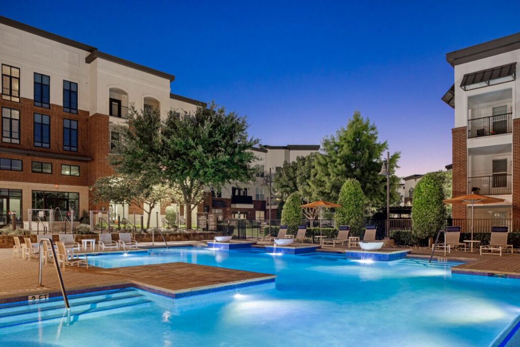 A modern apartment complex surrounds a lit outdoor swimming pool at dusk, with lounge chairs, umbrellas, and lush landscaping creating a relaxing atmosphere.