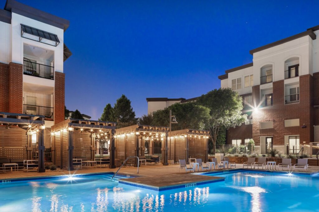 Outdoor swimming pool with bright lights, surrounded by lounge chairs, pergolas, and modern multi-story apartment buildings at dusk.