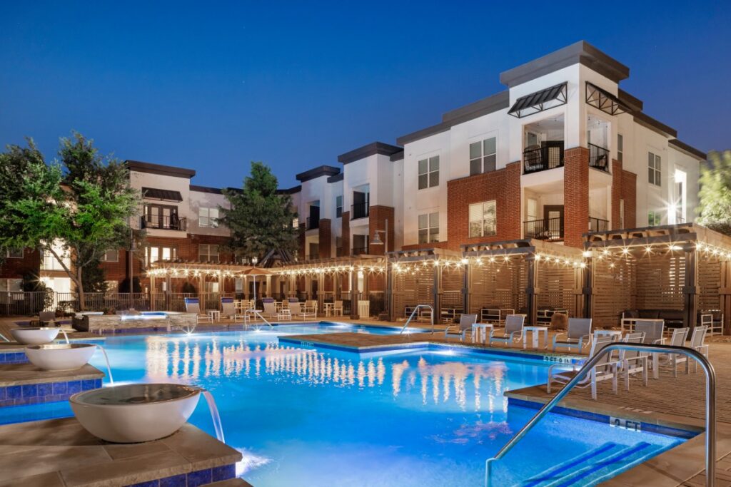 Modern apartment complex at dusk with illuminated swimming pool, lounge chairs, string lights, and landscaped greenery. The building features balconies and large windows, creating a welcoming, resort-like atmosphere.