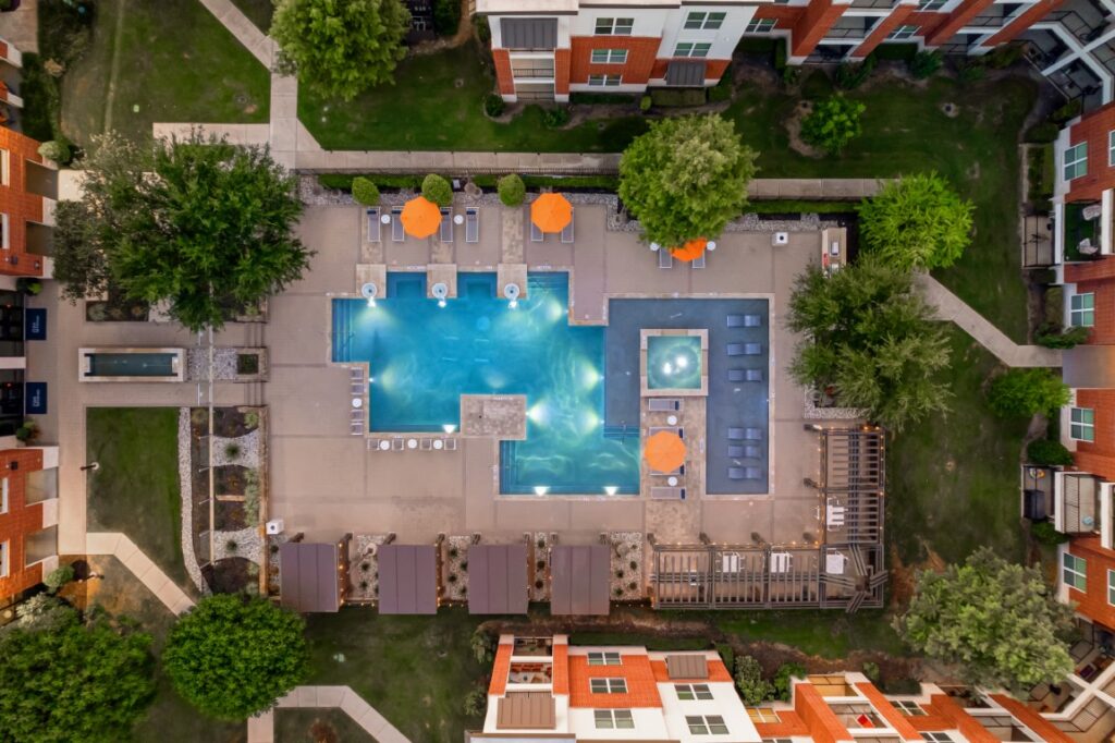 Aerial view of an apartment complex pool area with lounge chairs, orange umbrellas, hot tub, and surrounding green landscaping.