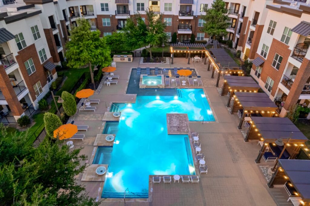 Aerial view of an apartment complex courtyard featuring a large, illuminated swimming pool, hot tub, lounge chairs, orange umbrellas, cabanas, string lights, and surrounding apartments.