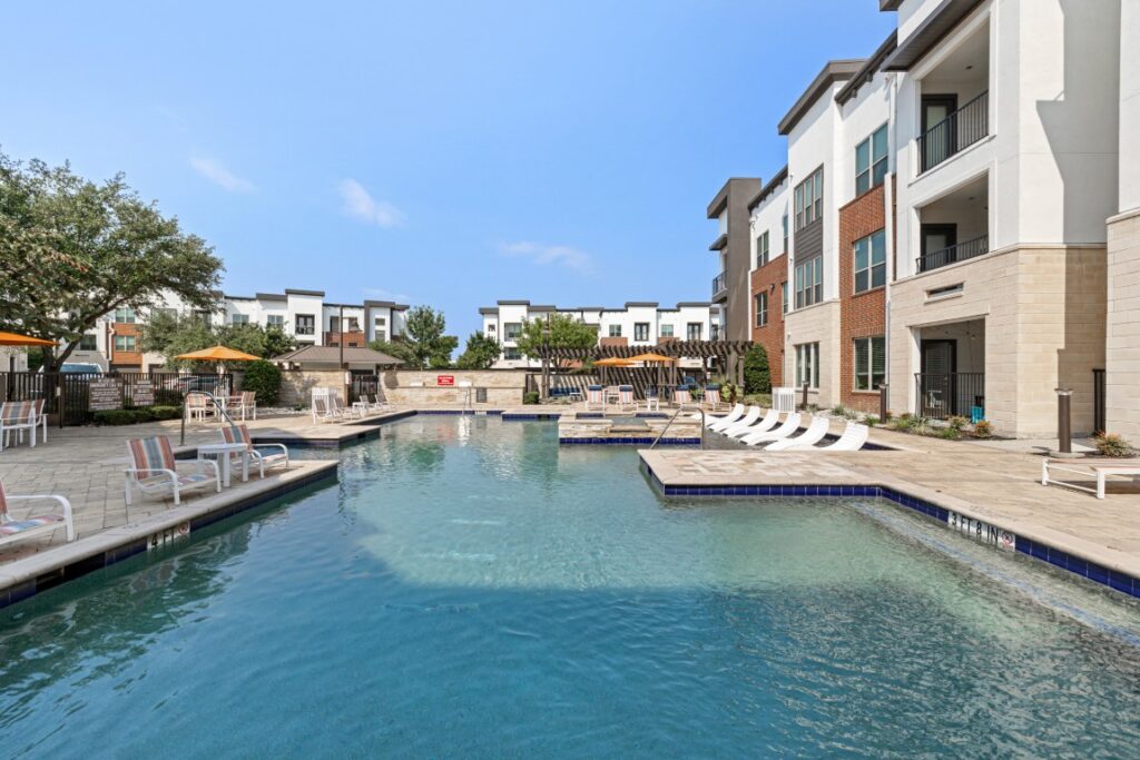 Modern apartment complex pool area with lounge chairs, umbrellas, and nearby buildings under a clear blue sky. Trees and seating areas surround the pool, creating a relaxing outdoor space.