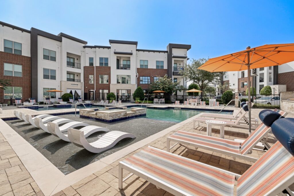 Modern apartment complex pool with submerged lounge chairs, striped poolside loungers, orange umbrellas, and three-story buildings in the background under a clear blue sky.