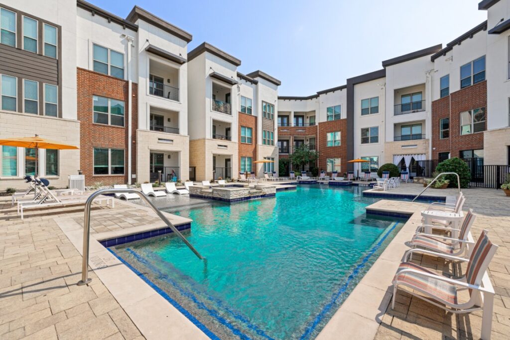 Modern apartment complex with a swimming pool, lounge chairs, umbrellas, and poolside seating. The buildings are white with brown brick accents and have balconies overlooking the pool area.