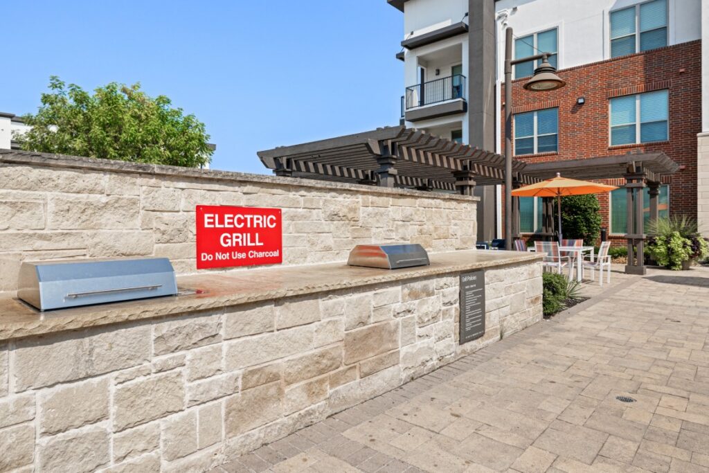 Two electric grills built into a stone counter on a patio, with a red sign that says ELECTRIC GRILL Do Not Use Charcoal. Outdoor tables with orange umbrellas and apartment buildings are visible in the background.
