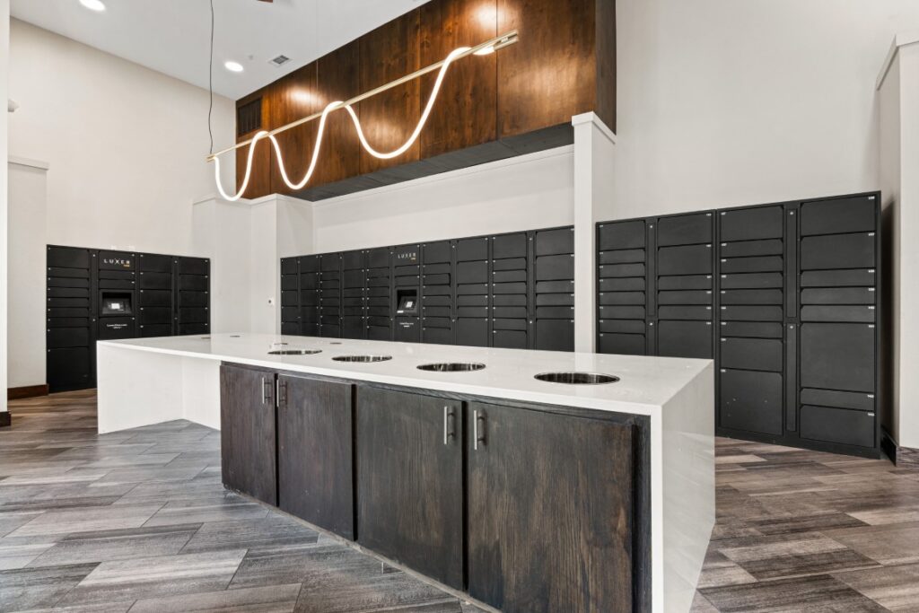 Modern mailroom with dark parcel lockers along the walls, a central countertop with four cutout holes, wood cabinets below, and a unique wavy pendant light above. The room features wood-look tile flooring and high ceilings.
