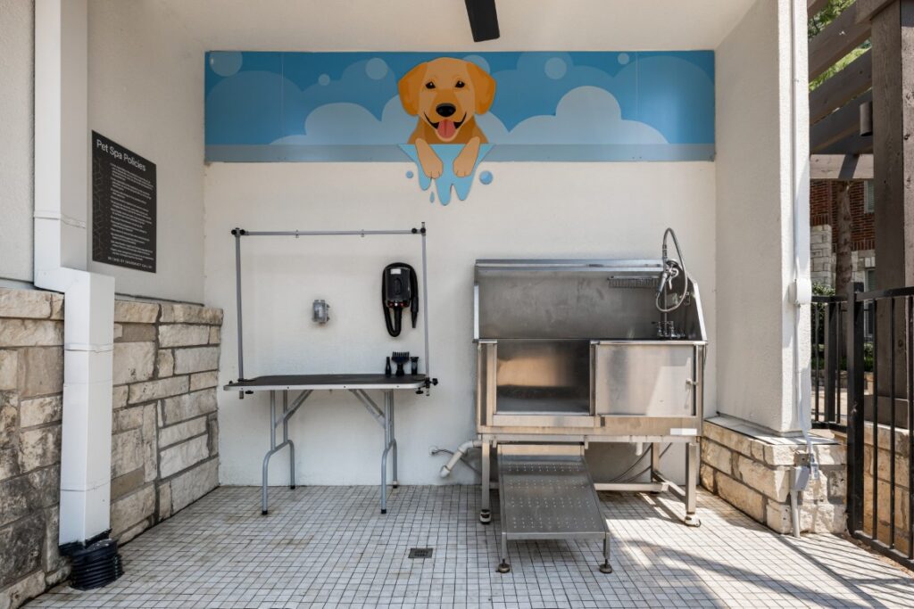 Outdoor pet washing station with a stainless steel tub, grooming table, soap dispenser, and mural of a cartoon yellow dog above. The area is clean, tiled, and partially enclosed with stone and metal accents.
